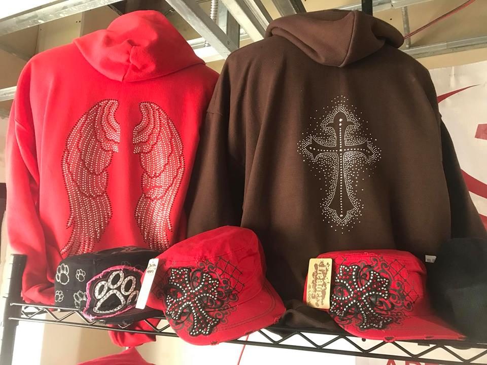 Red and brown hoodies with rhinestone wing and cross designs alongside matching caps